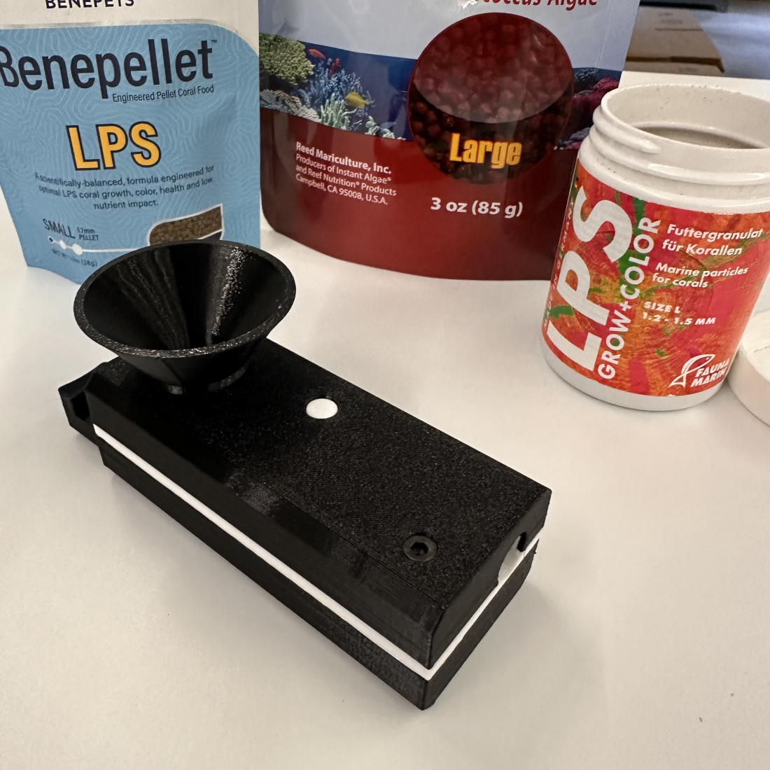 Black aquarium filter with fish food packaging in the background on a white surface. Avast Marine MATEY funnel and food